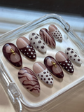 A set of 10 press-on false nails displayed in a clear plastic case. The nails feature a mix of patterns including solid deep brown, brown with white polka dots,  white with brown polka dots, and light brown with brown zebra stripes.