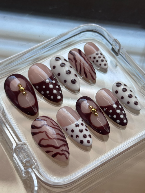 A set of 10 press-on false nails displayed in a clear plastic case. The nails feature a mix of patterns including solid deep brown, brown with white polka dots,  white with brown polka dots, and light brown with brown zebra stripes.