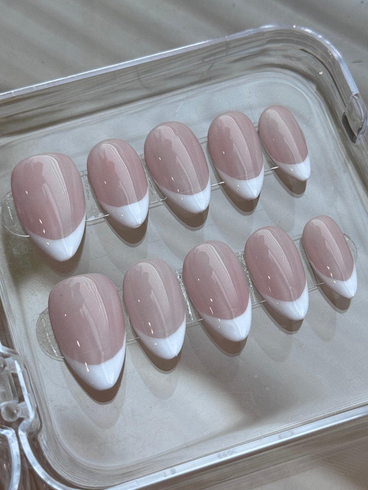A set of ten press-on nails with a classic French manicure design, featuring a dusty pink base and white tips, displayed inside a clear plastic case.
