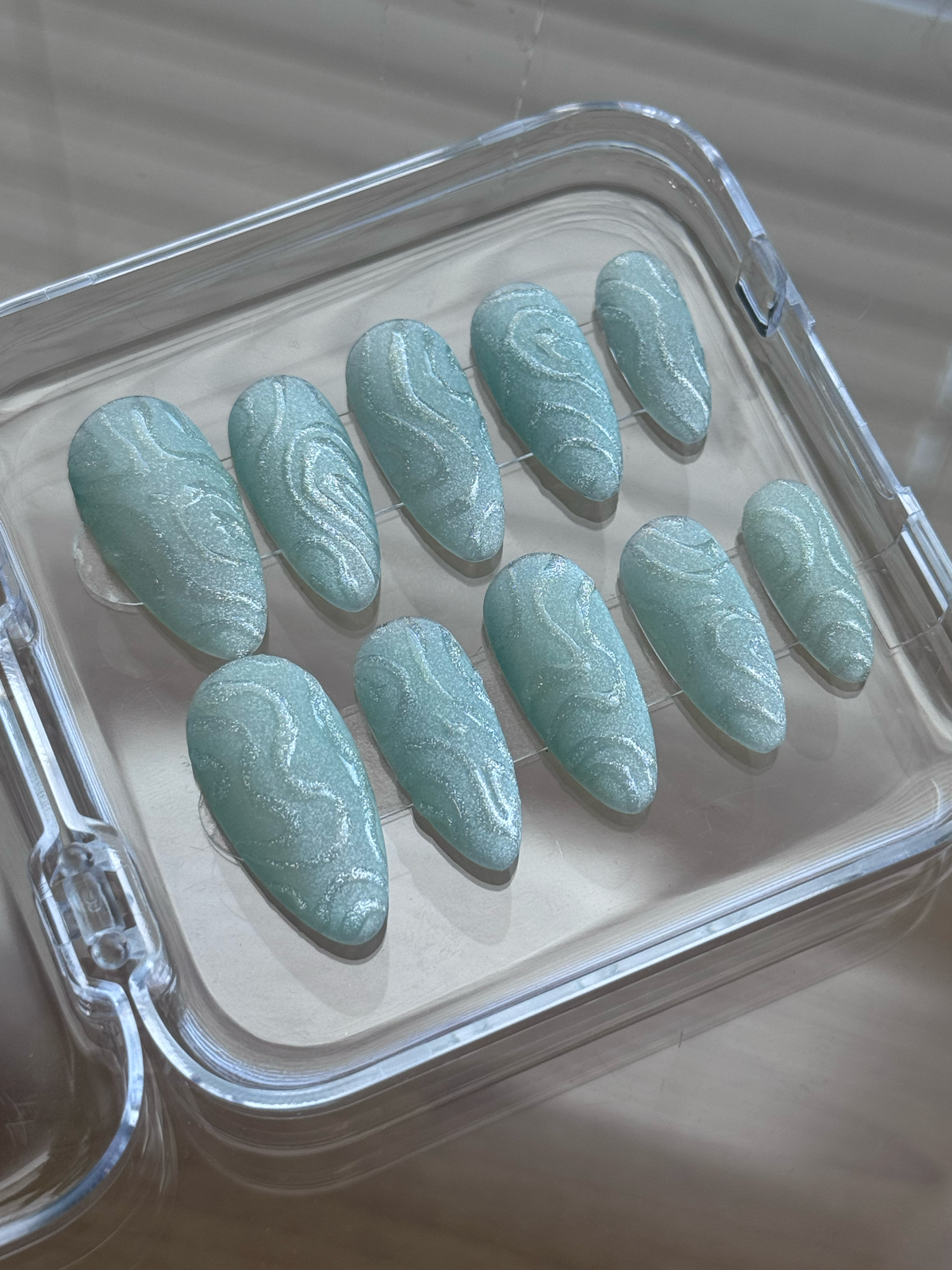 A set of ten almond-shaped press-on nails with a light green glitter finish and a clear wavy pattern, displayed inside a clear plastic case.

