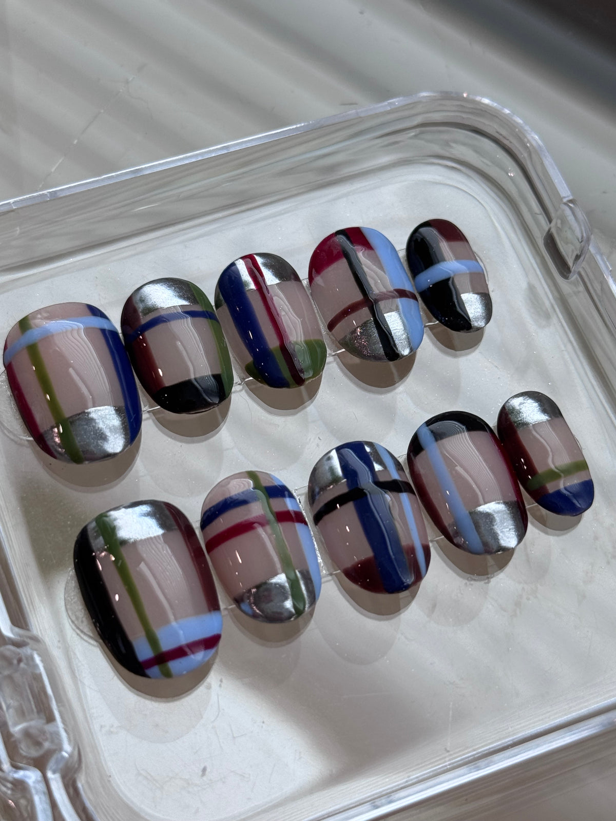 A set of 10 press-on nails with a glossy finish, featuring a striped pattern in black, silver, blue, red, green, and beige, arranged in a clear plastic tray.