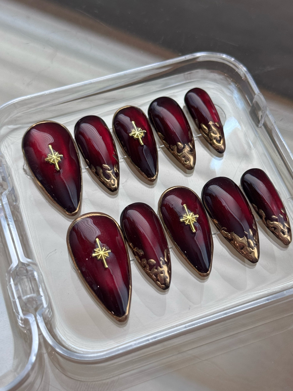 A set of glossy dark red press-on nails with gold metallic accents and star motifs, displayed in a clear plastic case.